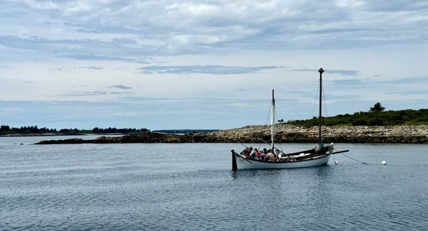 From a distance, a sailboat with white sails floats on water under a gray-blue sky.